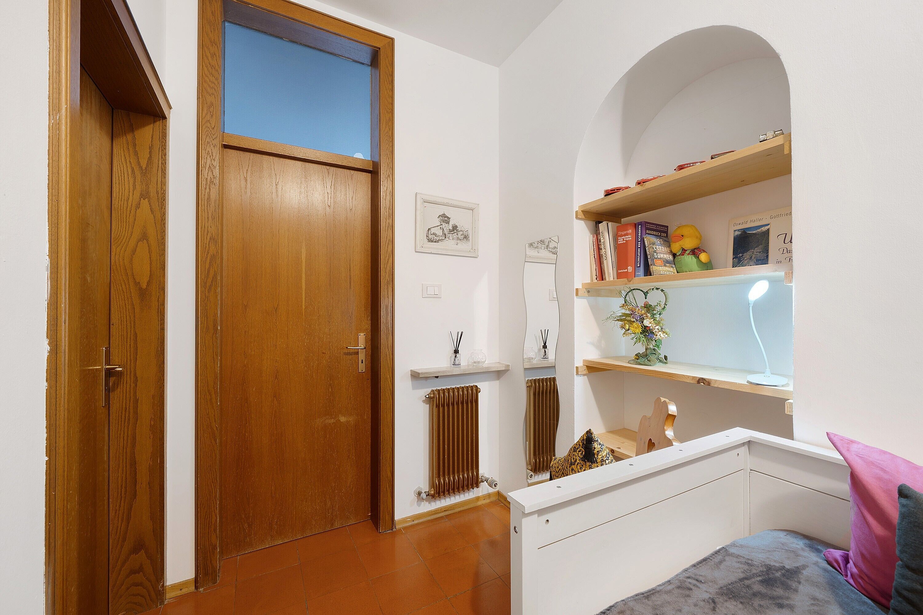 Ein Schlafzimmer mit Holztür, Heizkörper, einem Regal mit Büchern und einem Stofftier sowie einem Bett mit Kissen.