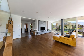 Una sala de estar moderna con un televisor de pantalla plana grande, piso de madera, un sofá amarillo y un ventanal corredero que da a un área exterior.