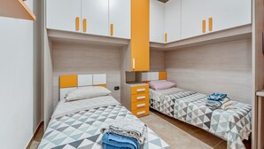 A room with two beds, each with a geometric-patterned bedspread, a wooden headboard, and a white wall. There are orange cabinets above the beds and a wooden bench in the corner. - Apartment 'Casa Rustica' with Private Terrace, Wi-Fi and Air Conditioning (Gonnesa)