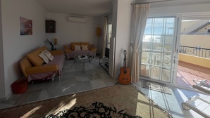 Una sala de estar con sofá, mesa de centro y una guitarra. Tiene un balcón con barandilla y vistas al exterior.