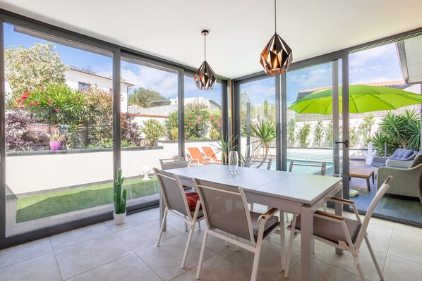 A dining area with a table and chairs, large windows, and a view of a swimming pool and garden.