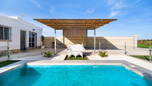 A modern outdoor pool area with a pergola, a white outdoor sofa, and a cacti plant. - Casa la Viña with pool, Wi-Fi, and air conditioning (Chiclana de la Frontera)