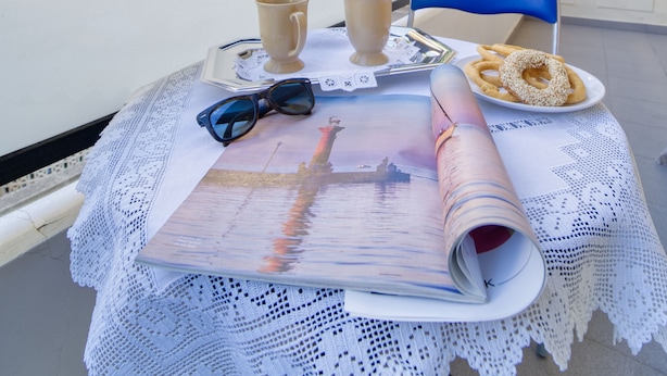 A balcony with a table set for two, featuring a lace tablecloth, a plate with onion rings, a cup, a pair of sunglasses, and a book. - ParkApart Rhodes Town (Rhodes)