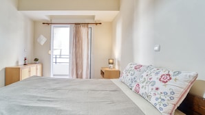 A bedroom with a large bed, a wooden dresser, a nightstand with a lamp, and a window with curtains. - ParkApart Rhodes Town (Rhodes)