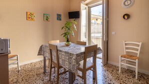 A dining area with a tiled floor, a table with a patterned tablecloth, chairs, a potted plant, and a television on the wall.