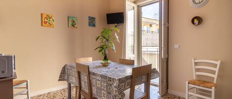 A dining area with a tiled floor, a table with a patterned tablecloth, chairs, a potted plant, and a television on the wall.