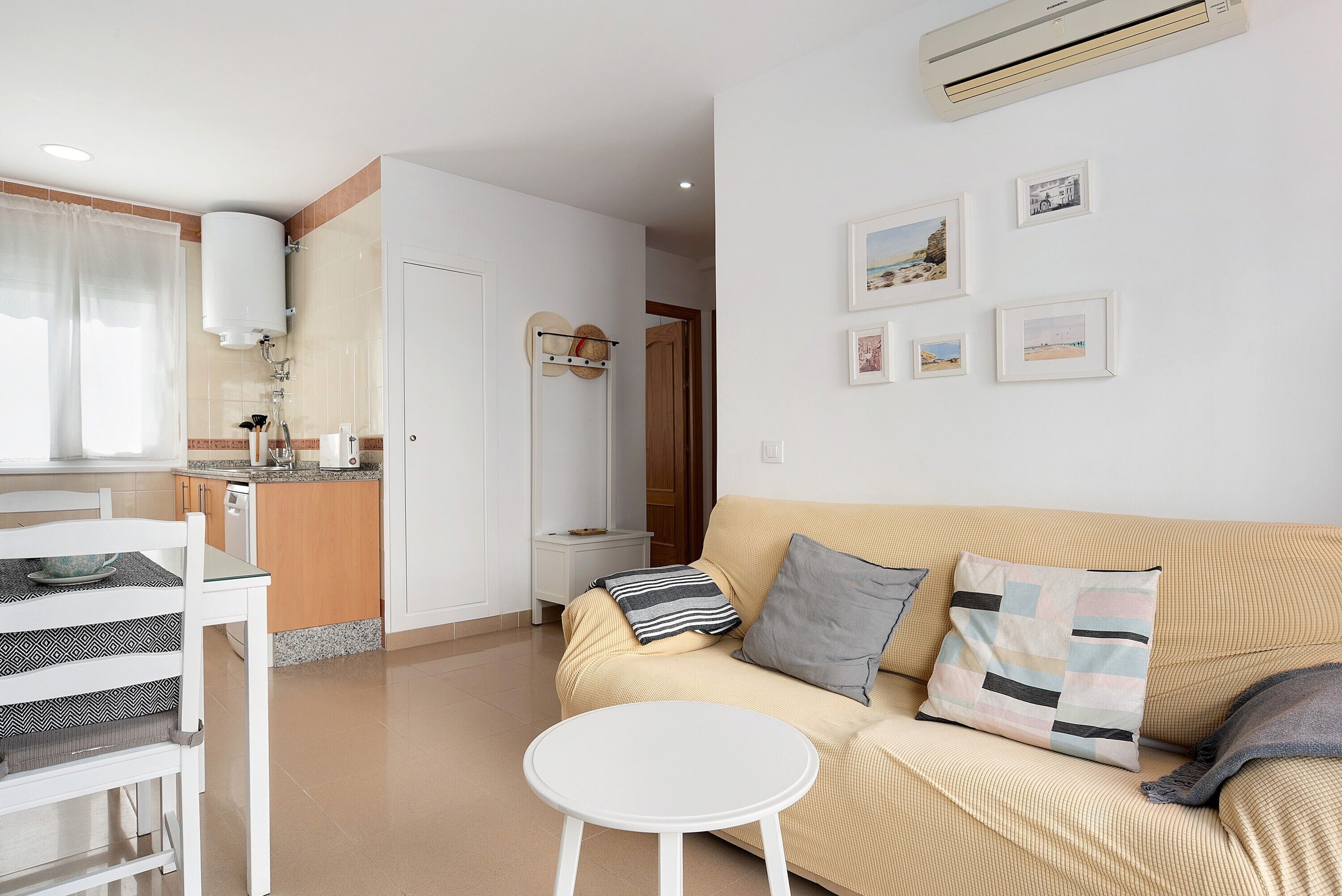 Un salón con un sofá color crema, una mesita redonda y una silla blanca. Incluye una zona de cocina con encimera y microondas.