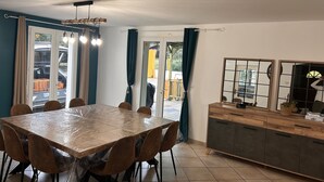 A dining area with a large table and chairs, a kitchen area with cabinets, and a view of the outside through a glass door.