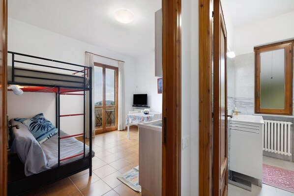 Una cama individual con televisor al fondo, una kitchenette pequeña y una puerta corrediza de vidrio que da a un espacio exterior.