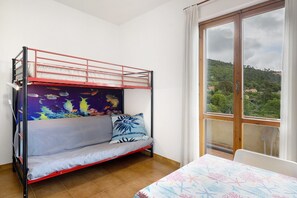 Una cama litera con un mural, un sofá con un cojín, una cama con una colcha estampada y vistas a un paisaje verde a través de una ventana.
