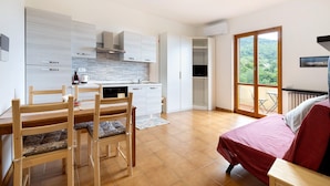 A modern kitchen with white cabinetry, a dining area with wooden chairs, a bed with a red cover, and a balcony with a view of greenery. - Apartment 'Elena E Joerg 2' with Mountain View, Shared Garden and Wi-Fi (Vezzi Portio)