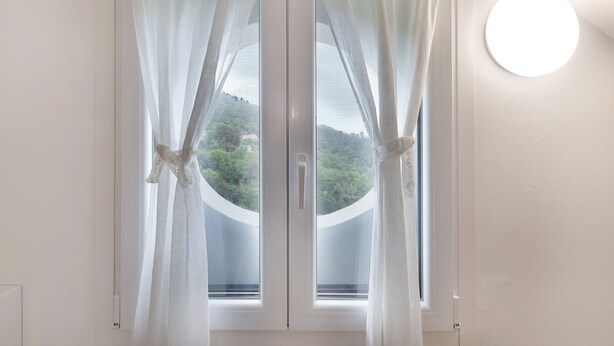A window with sheer curtains, a round wall light, and a view of greenery outside. - Apartment 'Elena E Joerg 2' with Mountain View, Shared Garden and Wi-Fi (Vezzi Portio)