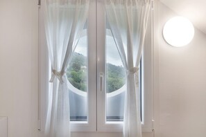 A window with sheer curtains, a round wall light, and a view of greenery outside. - Apartment 'Elena E Joerg 2' with Mountain View, Shared Garden and Wi-Fi (Vezzi Portio)