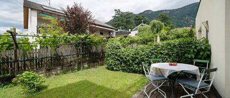 Eine Holzterrasse mit Tisch und Stühlen, ein eingezäunter Garten und im Hintergrund eine Aussicht auf Häuser und Berge.