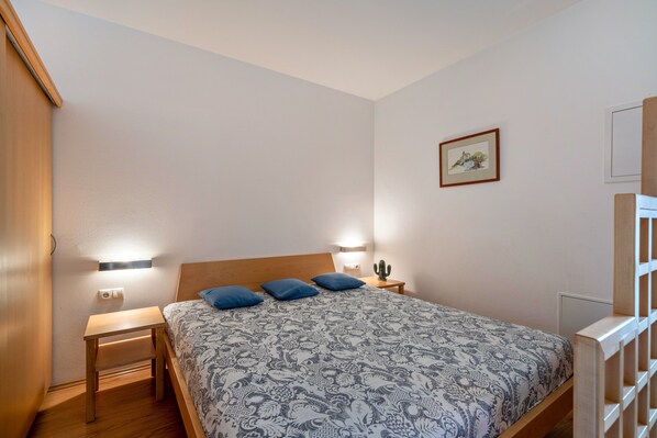 Une chambre à coucher avec un lit, des tables de chevet, une commode, une lampe fixée au mur, un cactus et un tableau encadré.