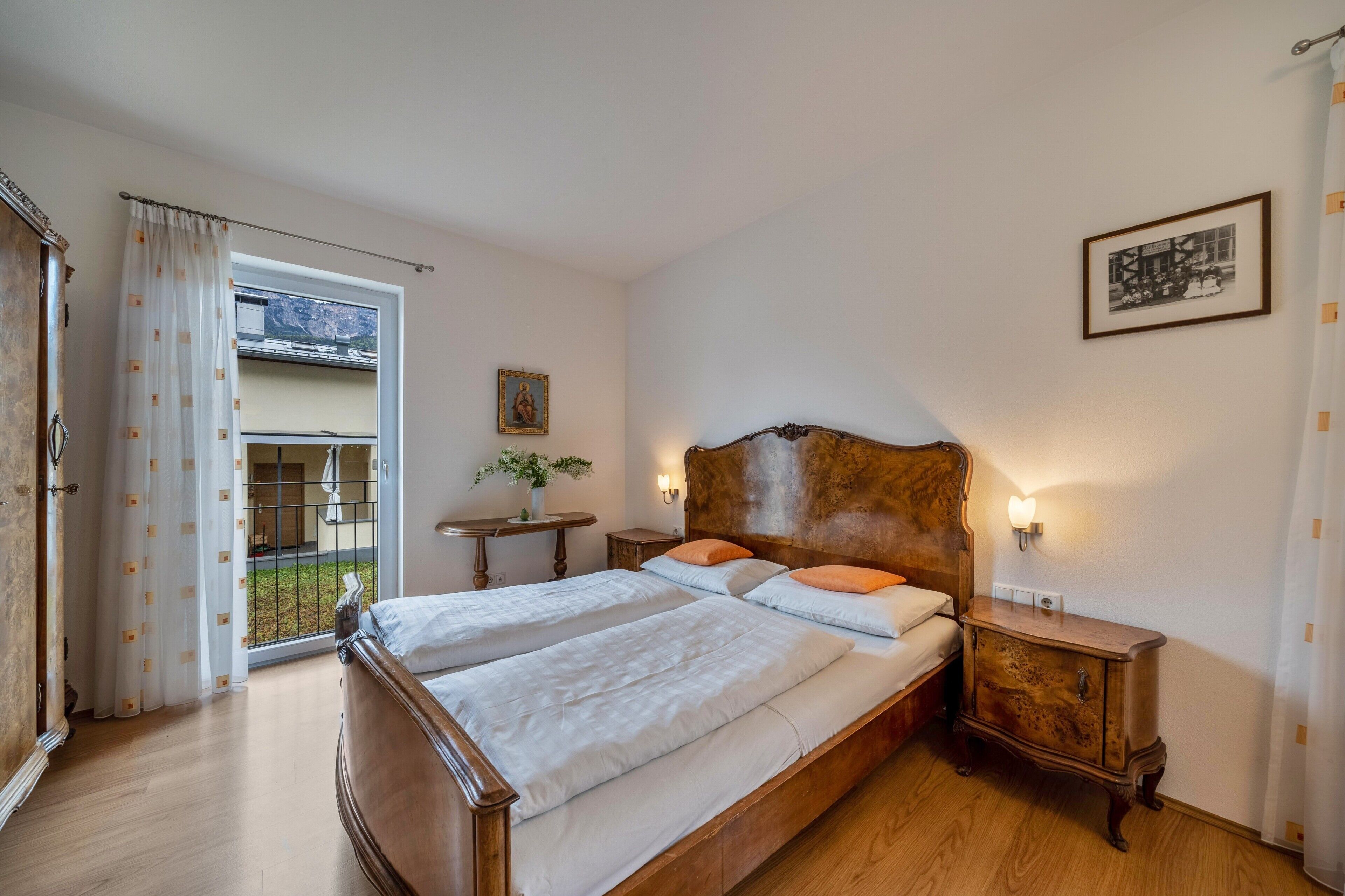 Une chambre à coucher avec un lit en bois, deux tables de chevet, une commode, une fenêtre avec des rideaux, une petite table sur laquelle est posé un vase, et un tableau encadré accroché au mur.