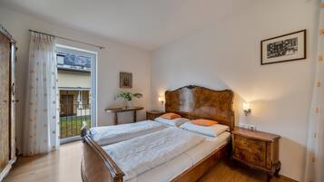 Une chambre à coucher avec un lit en bois, deux tables de chevet, une commode, une fenêtre avec des rideaux, une petite table sur laquelle est posé un vase, et un tableau encadré accroché au mur.