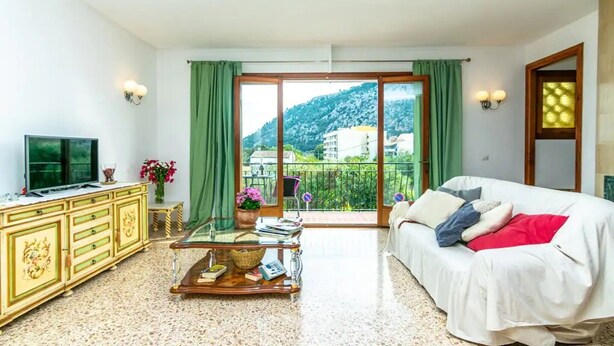 A room with a bed, a TV, a table with books, a potted plant, and a view of a mountain. - Holiday Home 'Puig' with Mountain View, Wi-Fi and Air Conditioning (Pollensa)