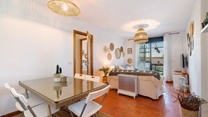 Un comedor con una mesa de vidrio y sillas blancas, una cama con edredón blanco, un radiador y un televisor sobre un mueble.