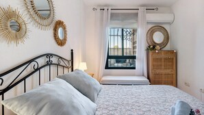 Un dormitorio con cama, cortinas en la ventana, espejo en la pared, mesita de noche con lámpara y cómoda.