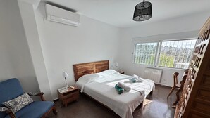 Quarto com cama, cadeira, mesa de cabeceira, abajur e janela com vista.