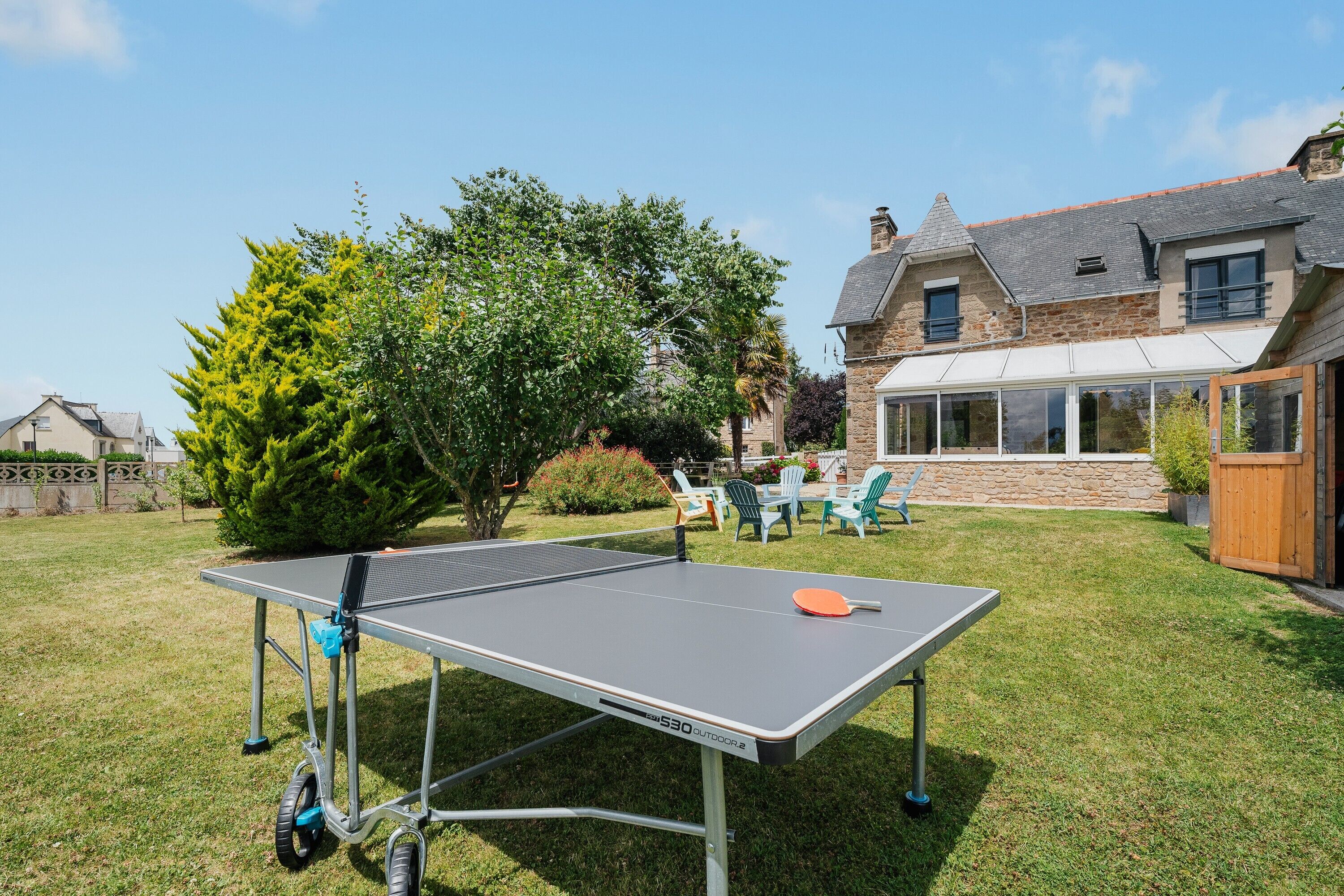 Une table de ping-pong installée à l’extérieur, dans un jardin, avec une maison en arrière-plan.