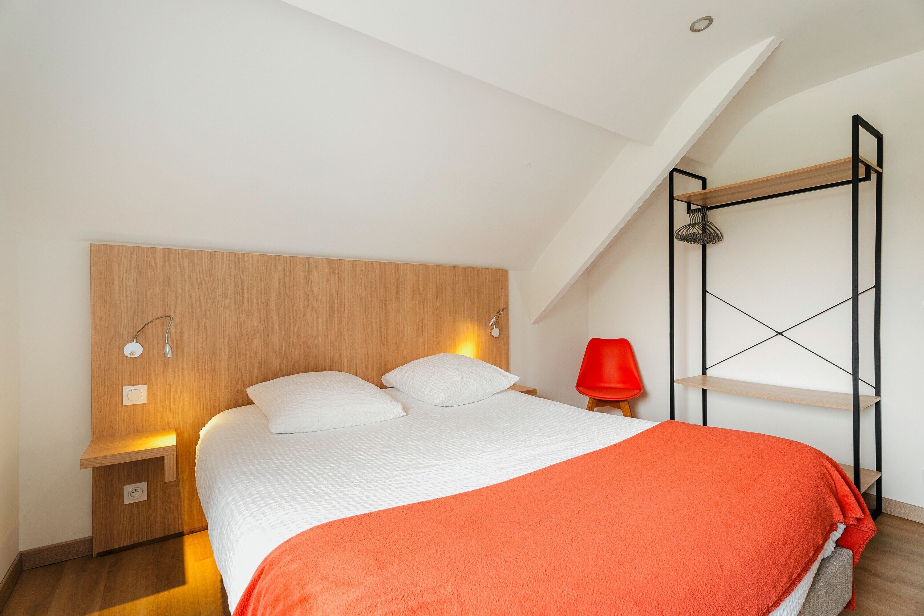 A bedroom with a bed, a nightstand, a red chair, a wooden headboard, and a black metal shelf.