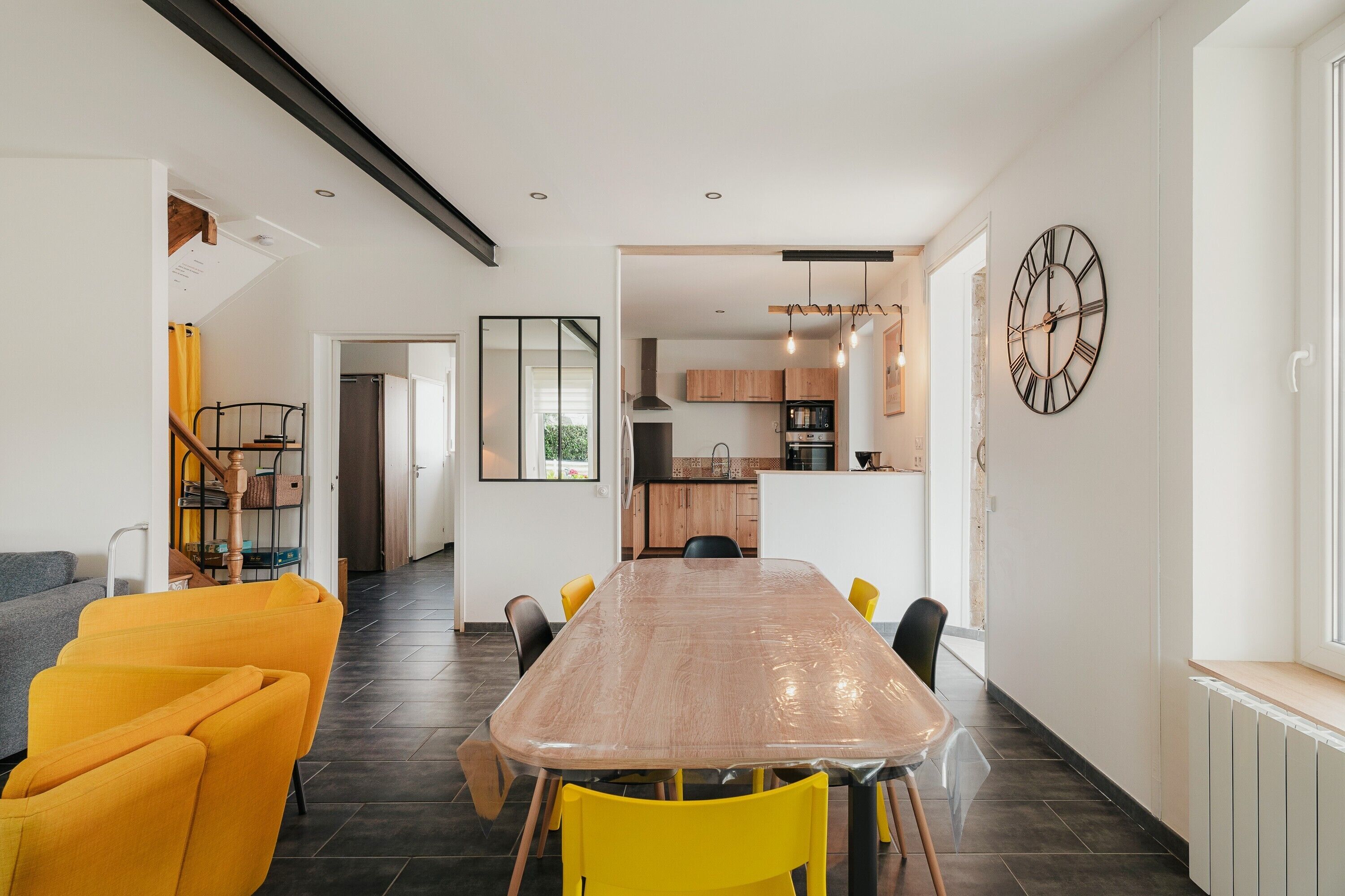 Une cuisine moderne dotée d’un grand îlot central, d’un coin repas avec une longue table et des chaises jaunes, et d’une horloge bien visible au mur.