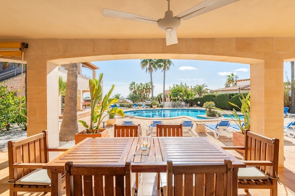 A covered outdoor seating area with a pool view, wooden furniture, and a ceiling fan.