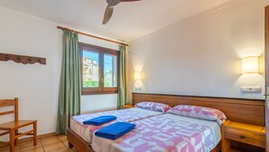 A room with two beds, a chair, a wooden nightstand, a ceiling fan, and a window with curtains.