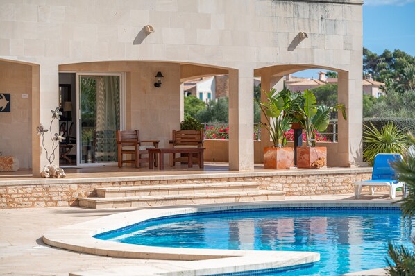 Un área junto a la piscina con un muro de piedra, una zona de estar cubierta con mesa y sillas, y vistas del paisaje circundante.