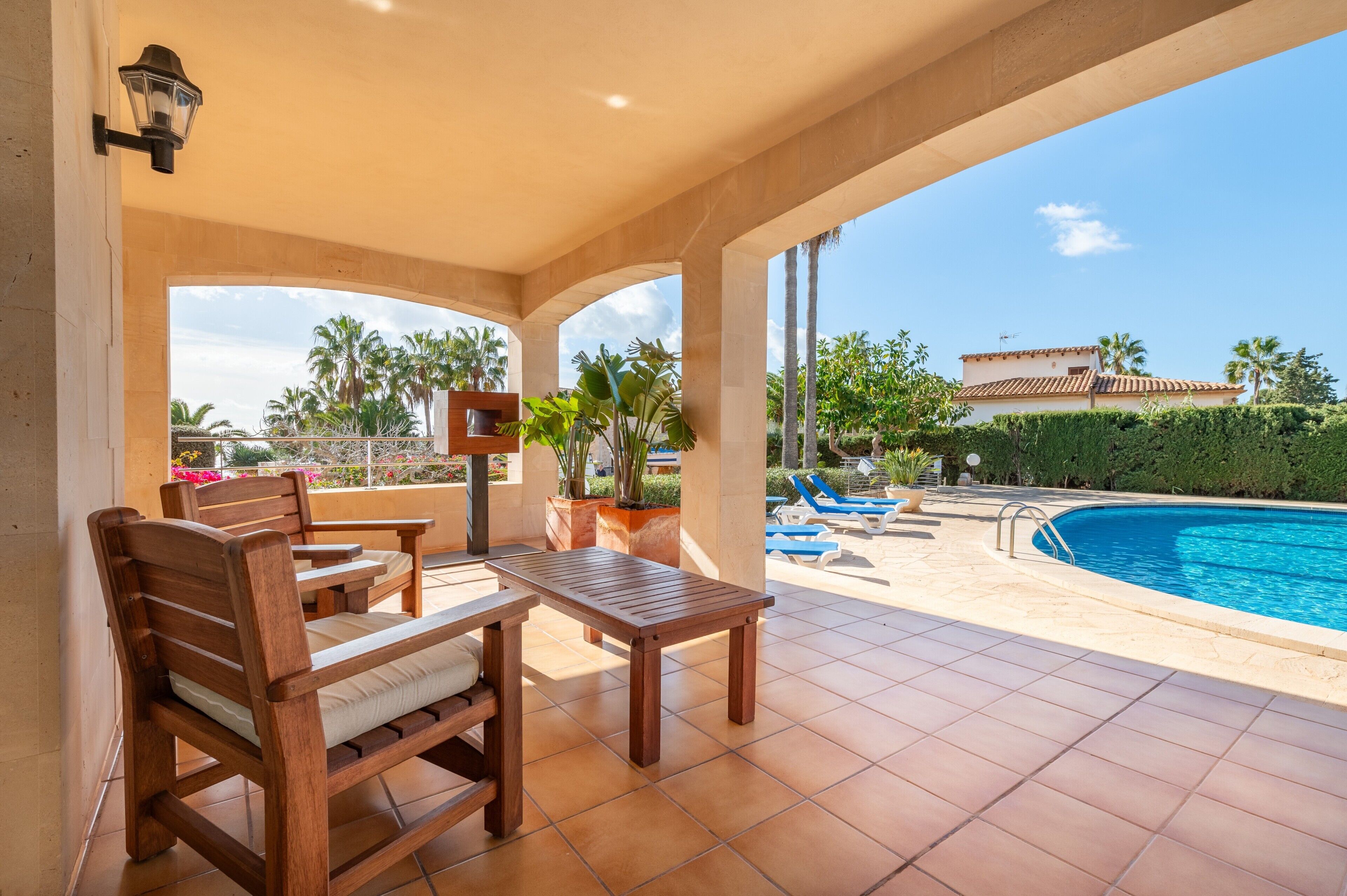 Eine überdachte Terrasse mit Holzmöbeln, einem Pool und Blick auf ein Haus und Palmen.