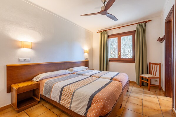 Un dormitorio con una cama de madera, dos mesitas de noche, un ventilador de techo, una ventana con cortinas y una silla.