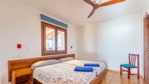 Ein Schlafzimmer mit Bett, einem Holznachttisch, einem Sessel, einem Deckenventilator, einem Fenster und einer Tür.