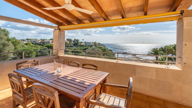 Ein Holztisch und Stühle auf einem Balkon mit Blick auf das Meer und entfernte Gebäude.