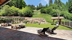 Une terrasse carrelée en pierre, dotée de meubles en bois et offrant une vue sur une colline boisée.