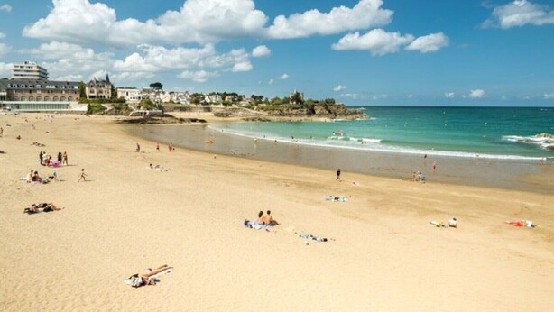 A beach with people sunbathing and swimming, a clear sky, and buildings in the background.