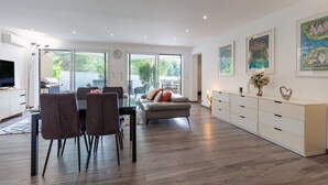 A spacious living area with a dining table, chairs, a sofa, and a TV. There are large windows, a white cabinet, and artwork on the walls.