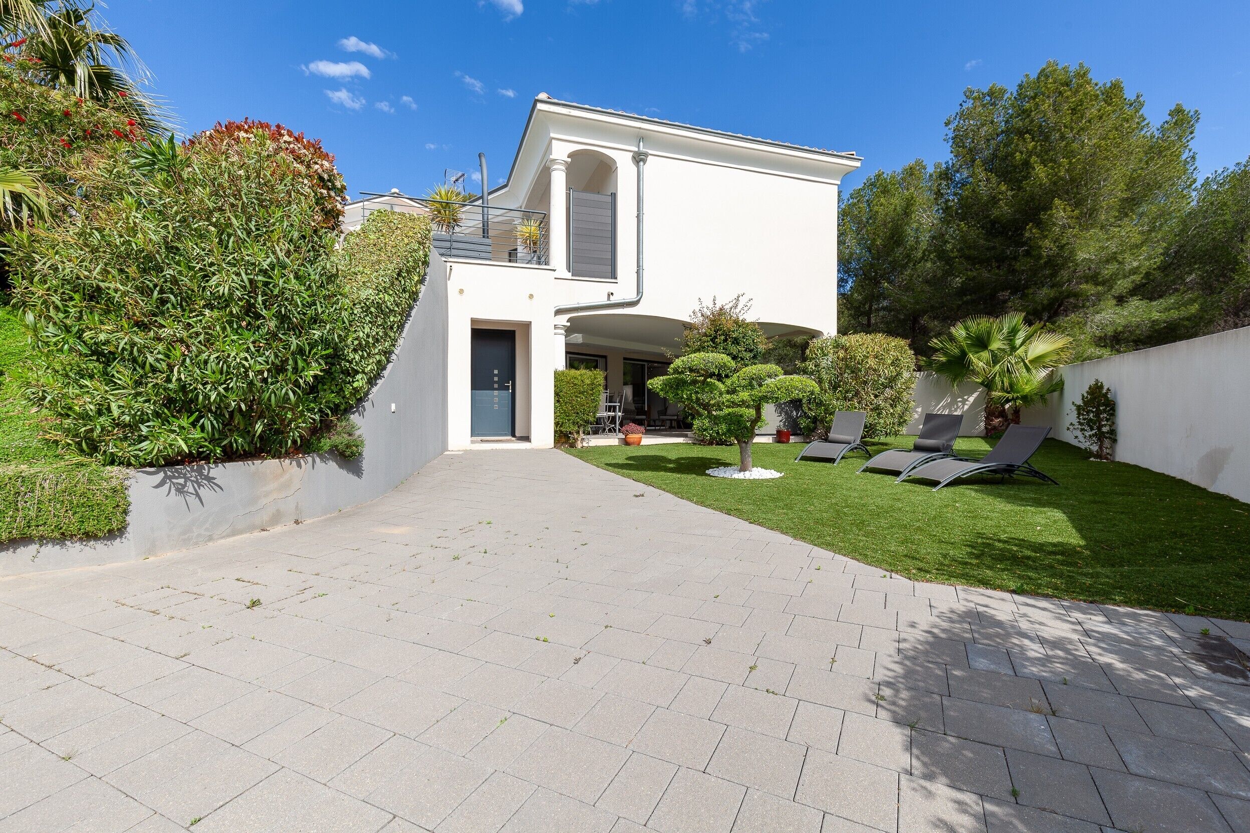 Una casa moderna de dos pisos con camino de entrada pavimentado, un pequeño jardín con una palmera y un balcón con barandilla.