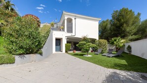 A modern two-story house with a paved driveway, a small garden with a palm tree, and a balcony with a railing.