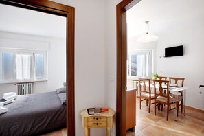 Una habitación con cama, una mesita con revistas, un comedor con sillas, un televisor en la pared y una ventana con cortinas.