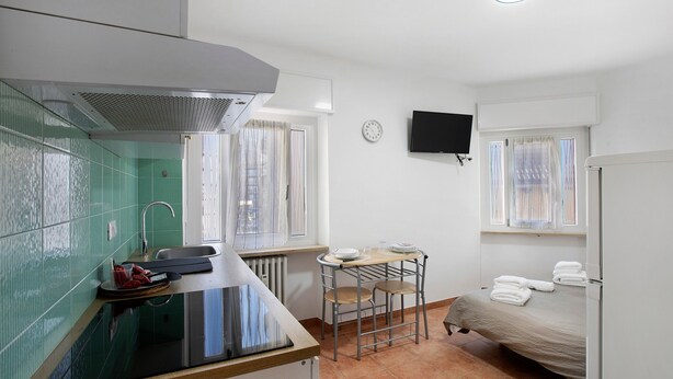 Ein kleines Zimmer mit Küchenzeile, einem Bett, einem Fernseher und einem Fenster mit Vorhängen.
