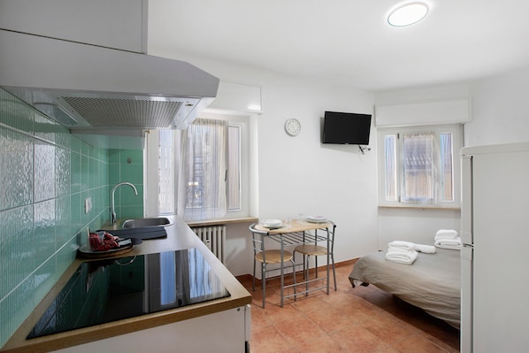 A compact room with a kitchenette, a bed, a TV, and a window with curtains.