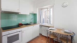 Eine kleine Küche mit weißen Schränken, einer grünen Spritzschutzleiste, einem Fenster mit Vorhängen und einer Wanduhr.