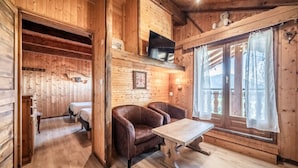 A wooden interior with a bed, two leather chairs, a table, a TV, and a window with curtains.
