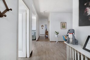 Ein Flur mit Holzboden, weißen Wänden, einem kleinen Tisch mit einer Pflanze, einem gerahmten Bild und einer grau farbenen Hundestatue.
