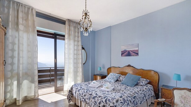 A bedroom with a bed, a chandelier, a view of the outdoors, and a painting on the wall. - Bella Vista Apartment in Premeno with Lake Maggiore View (Premeno)