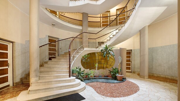 Un interior majestuós amb una escala corba, una font central i plantes en testos.