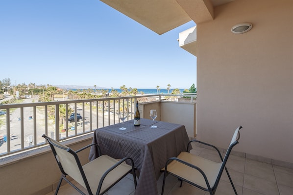 A balcony with a table and chairs, overlooking a parking area and a view of the sea.