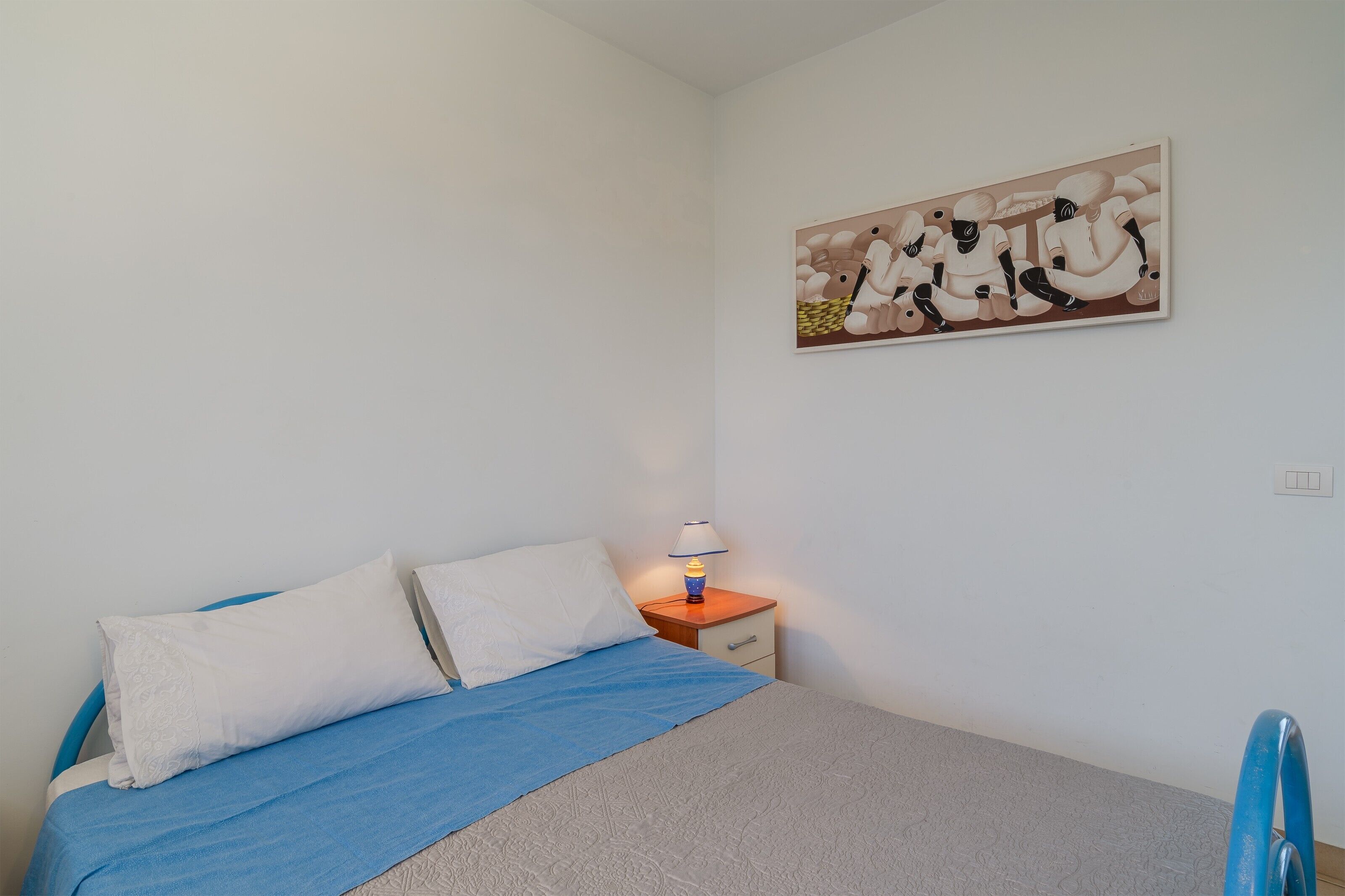 A bedroom with a bed, pillows, a bedside table with a lamp, and a wall-mounted painting.